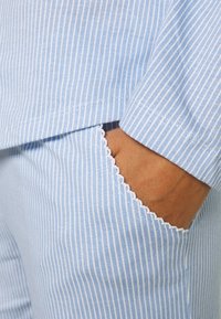 Mėlynos ir baltos juostelės audinys su išgaubtu kišenės krašto dekoru. Jo tekstūra glotni, tinkama kasdieniam ar laisvalaikio drabužių stiliui.