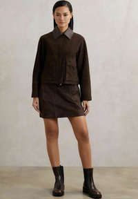 Ruda suede striukė su pilku apykakle, derinama su atitinkančia rudą suede mini sijonu. Juodi aulinukai užbaigia aprangą.