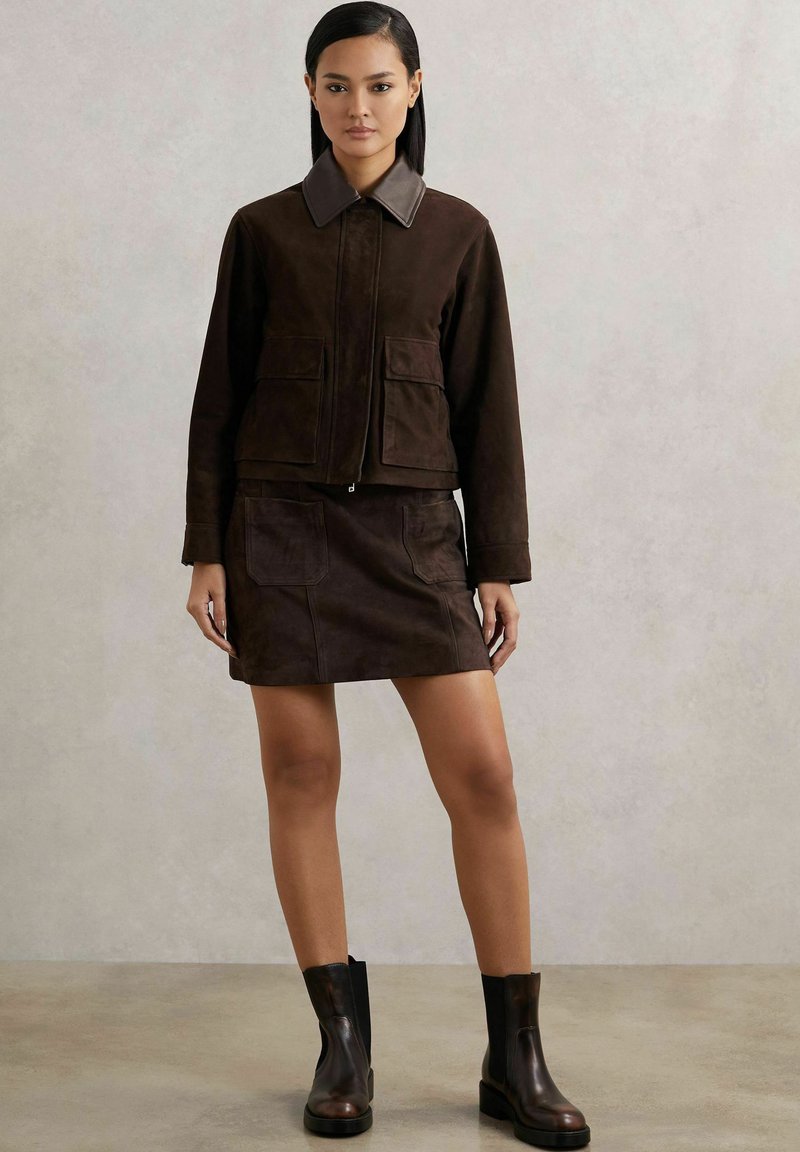 Ruda suede striukė su pilku apykakle, derinama su atitinkančia rudą suede mini sijonu. Juodi aulinukai užbaigia aprangą.