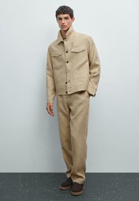 Beige, strukturierte Jacke mit zwei Brusttaschen und Knopfverschluss, kombiniert mit passenden, lockeren Hosen und braunen Schuhen auf einer dunklen Oberfläche.