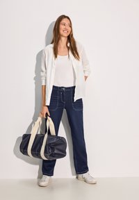 Pantaloni in denim blu scuro con cuciture a contrasto, abbinati a una canotta bianca e a una camicia leggera bianca con bottoni; tiene una borsa blu navy.