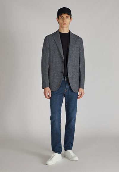 Grau strukturierte Blazer mit einem einzelnen Knopf, getragen über einem schwarzen Hemd, kombiniert mit dunkelblauen Jeans und weißen Sneakers. Eine schwarze Mütze rundet den Look ab.