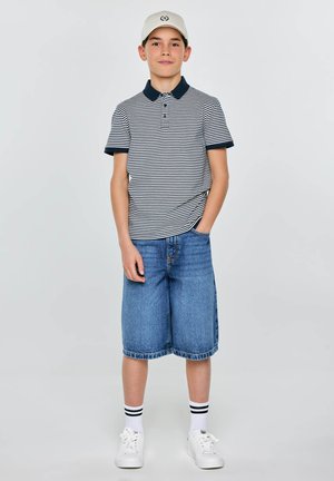 Chłopiec stoi z jedną ręką w kieszeni, ubrany w białą czapkę, pasiastą koszulkę polo, dżinsowe szorty do kolan, białe skarpetki i trampki na jednolitym tle.