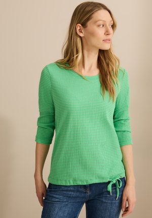 Haut long vert texturé avec un col rond, présentant un détail de lien sur le côté et un ourlet côtelé. Associé à un jean en denim bleu.