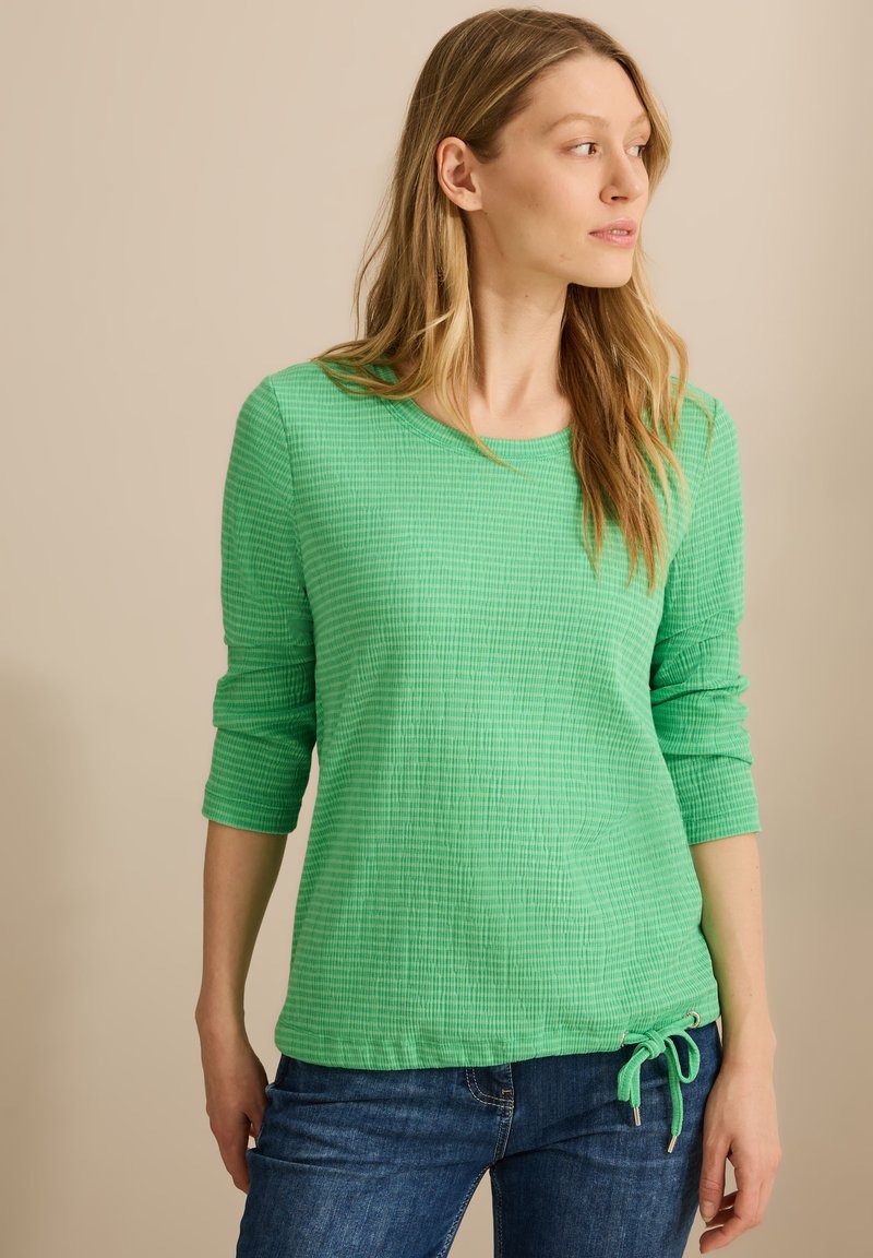 Maglietta verde a maniche lunghe, con colletto rotondo, caratterizzata da un dettagli di nodo laterale e orlo a coste. Abbinata a jeans in denim blu.