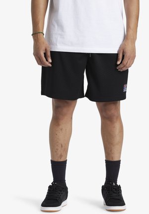 Pantaloni corti in rete nera con cintura elastica, dotati di tasche laterali e un piccolo logo ricamato. Indossati con scarpe da ginnastica nere e calze.