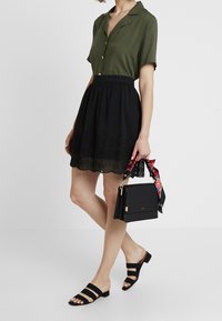 Olivgrön skjorta med korta ärmar och knappar, tillsammans med en svart kjol med blommönster vid fållen. Svart handväska med blommig detalj.