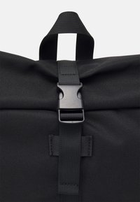 Sac en tissu noir avec une poignée de transport supérieure et une sangle réglable maintenue par une boucle de déverrouillage latérale en plastique noir.
