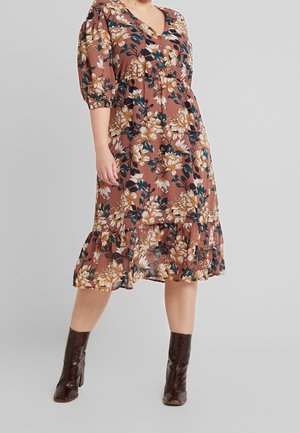 Femme portant une robe midi marron à fleurs avec des manches bouffantes et des bottines marron foncé à talons sur un fond gris uni.
