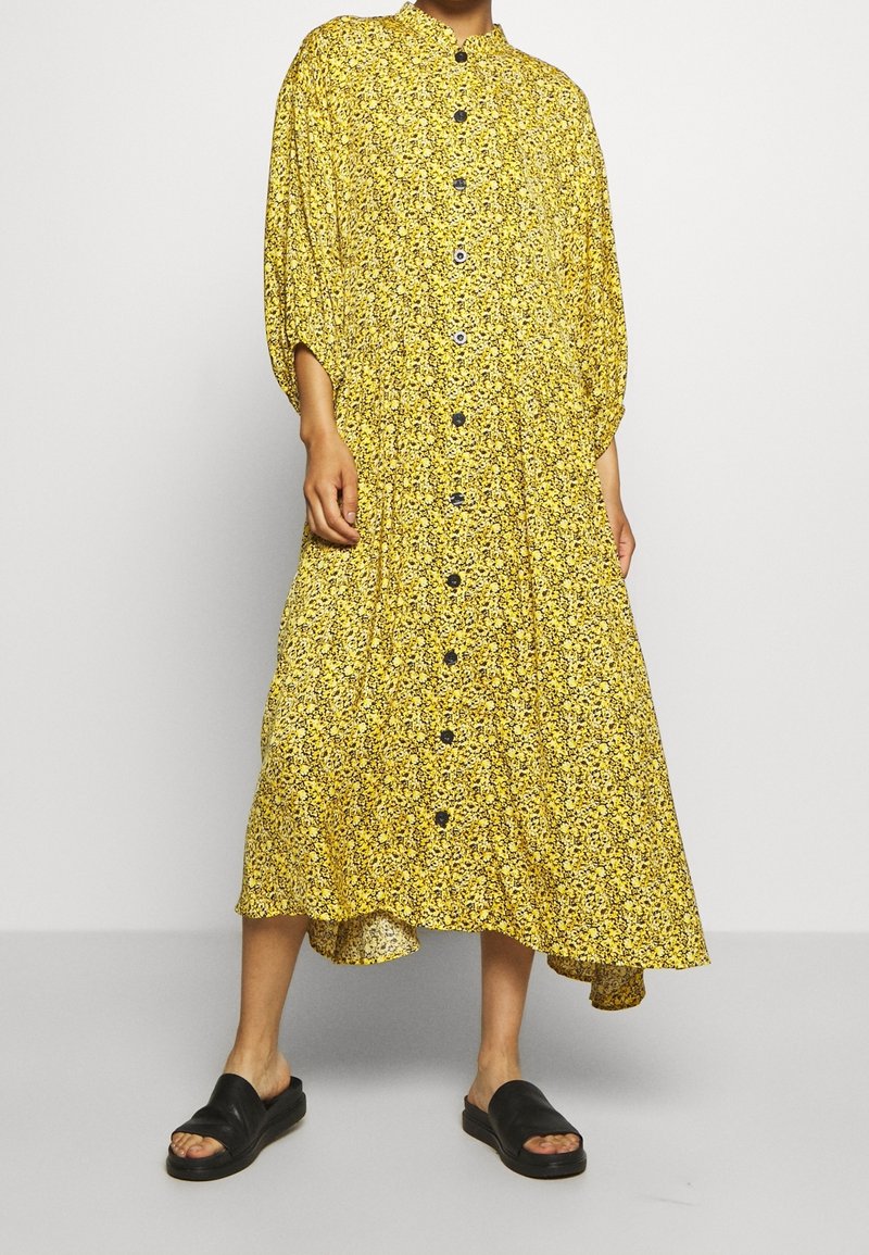 Robe midi à fleurs jaune à boutons, avec un ourlet haut-bas, dotée d'un col mandarin et de boutons noirs, portée avec des sandales plates noires.