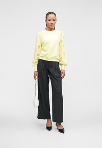 Kvinne med flettet hår har på seg en gul langermet topp med utskjæringer, svarte vide bukser, svarte hæler, og holder en hvit håndveske.