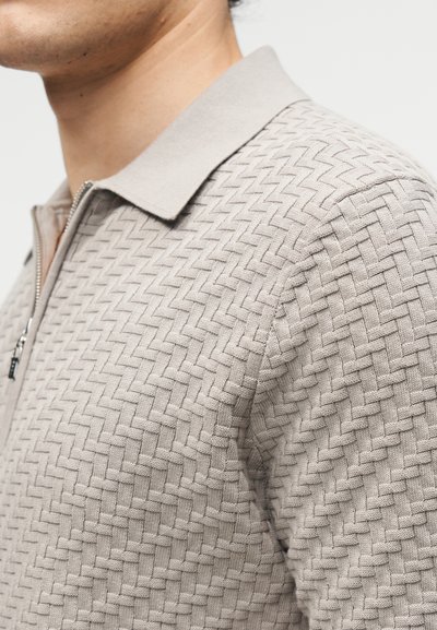 Άνδρας που φορά ένα ανοιχτό μπεζ πλεκτό πουλόβερ με φερμουάρ, γιακά και υφασμάτινο υφή με ζικ ζακ μοτίβο.