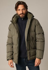 Doudoune vert olive avec une fermeture éclair à l'avant, une grande capuche et deux poches latérales. Pull texturé bleu marine en dessous. Coupe décontractée.