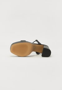 Chaussure à talon haut noire avec une tige en cuir lisse, des détails de sangle et une semelle en caoutchouc nervurée. Le talon est épais et de couleur beige.
