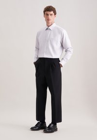 Camicia button-up grigia chiara con taschino sul petto, abbinata a pantaloni neri con pieghe e scarpe nere, su uno sfondo neutro.