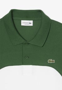 Vihreä ja valkoinen polo-paita, jossa on teksturoitu kangas, kaksi vihreää nappia ja pieni Lacoste-logo vasemmassa rinnassa. Säännöllinen istuvuus.