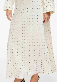 Robe longue couleur crème à texture lisse, ornée d'un motif de petits points noirs. Manches longues avec poignets et une fente latérale.