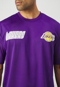 Mężczyzna ubrany w fioletową sportową siateczkową koszulkę Los Angeles Lakers z białym napisem i logo drużyny na piersi, zwrócony lekko w lewo.