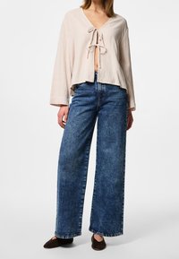 Cropped top beige chiaro con nodo frontale, tessuto testurizzato, abbinato a jeans in denim blu a vita alta e gamba larga, e scarpe scure a punta.