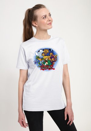 Kvinde med lyst brunt hår i hestehale iført en hvid t-shirt med farverige Yu-Gi-Oh! figurer og logo, som kigger opad mod siden.