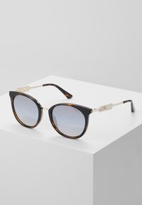 Occhiali da sole a forma di gatto in nero e tartaruga con lenti sfumate. Presentano il marchio dorato "GUESS" sugli accenti laterali.