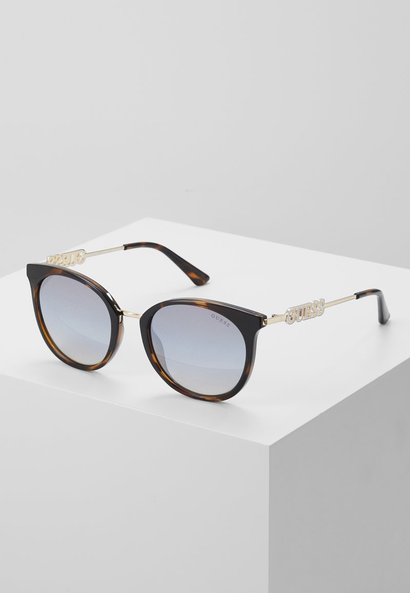 Occhiali da sole a forma di gatto in nero e tartaruga con lenti sfumate. Presentano il marchio dorato "GUESS" sugli accenti laterali.