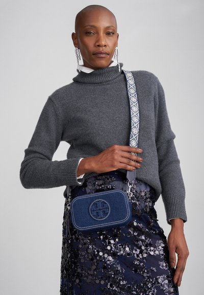 Mala de ombro em denim com fecho de zip, logótipo bordado e alça estampada. Suéter cinza com gola alta, saia com lantejoulas por baixo.