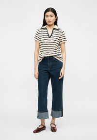 Νεαρή γυναίκα που φοράει μπλουζάκι με κοντά μανίκια σε άσπρες και μαύρες ρίγες, σκούρα μπλε τζιν με ρεβέρ και σκούρα κόκκινα παπούτσια Mary Jane σε λευκό φόντο.