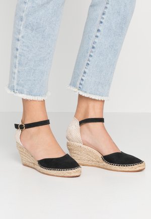 Pieds dans un jean court bleu clair portant des espadrilles compensées noires avec des talons en jute tressé et des brides de cheville, se tenant sur un sol blanc.