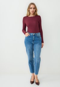 Burgunderrotes langärmliges Oberteil, taillierte Passform, kombiniert mit hochgeschnittenen blauen Jeans im geraden Schnitt, ergänzt durch dunkelbraune hohe Absätze.