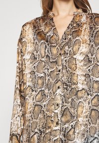 Långärmad skjorta i ormtryck med bruna, beige och svarta mönster. Har en krage och svarta knappar framåt. Sidenliknande textur.