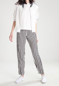 Bílá krajková bomber bunda s černým lemem, bílý tričko a černobílé pruhované kalhoty doplněné bílými teniskami.