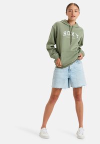Green hoodie with white "ROXY EST 1990" lettering, front pocket, paired with light blue denim shorts, and white sneakers. Casual outfit.