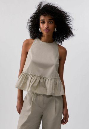 Donna con capelli ricci che indossa un top beige senza maniche con orlo arricciato e pantaloni beige coordinati su uno sfondo semplice.