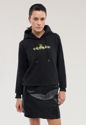 Felpa nera con logo frontale in verde neon, abbinata a una gonna aderente in pelle nera. Il tessuto sembra strutturato e morbido.