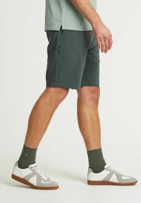 Pantaloni corti verde scuro con tasche laterali, indossati con scarpe bianche con dettagli grigi e calzini verdi. Tessuto liscio, vestibilità moderna, stile casual.