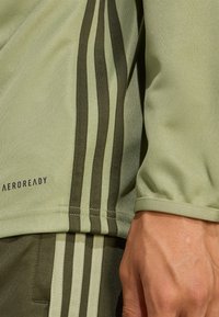Sportinė viršutinė dalis, pagaminta iš tekstūruoto šviesiai žalio audinio, su tamsiai žaliais dryžiais ir „AEROREADY“ logotipu, turinti apžvalgos rankoves.