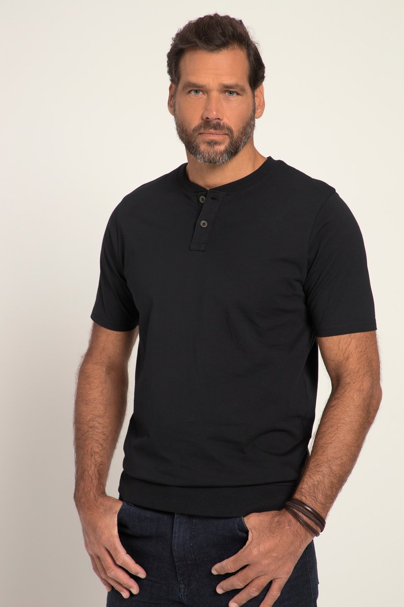 Hombre con cabello castaño corto y barba, vistiendo una camiseta henley negra de manga corta y pantalones oscuros sobre un fondo liso.