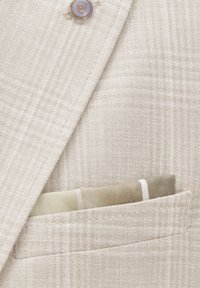 Hellbeige Blazer aus einem strukturierten Stoff, ausgestattet mit einem kleinen Taschentuch in einem sanften Grün- und Crememuster. Knopfdetails sichtbar.