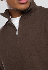 Brown zip-up sweater with a textured fabric, high collar, silver zipper, and white t-shirt beneath, featuring a close-up view of the neckline.