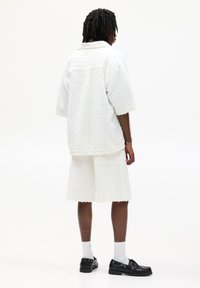 Hvid struktureret skjorte og matchende shorts, med en boxy pasform og frynset kanter. Båret med sorte sko og hvide sokker, set fra bagved.