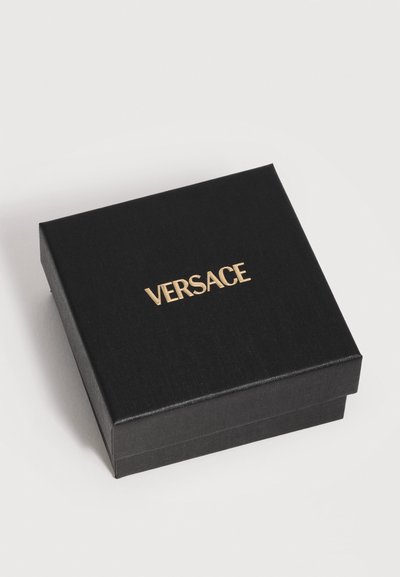 Boîte carrée noire avec le texte doré « VERSACE » au centre du couvercle, placée sur une surface plate gris clair.