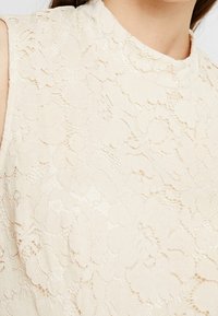 Närbild av en cremefärgad ärmlös topp med intrikat blommönstrad spetsöverdrag och hög krage på en person med rakt brunt hår.