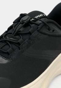 Fekete sportcipő elasztikus bungee fűzőkkel, csúszózáras megoldással, mintás felsőrésszel és fehér talppal, a nyelvén "TRANSPORT 2" felirattal.