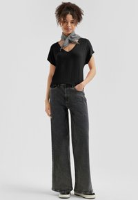 Top nero a costine con scollatura a V e maniche corte, abbinato a jeans grigi scuri a gamba ampia e una sciarpa grigia con motivo intorno al collo. Stile casual.