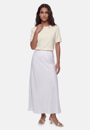 Mujer con el pelo ondulado lleva una camiseta crema de manga corta, falda larga blanca y sandalias beige de tacón bajo, de pie frente a un fondo liso.