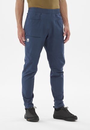 Homme portant un pantalon de randonnée bleu fuselé avec une poche zippée sur la cuisse et des chaussures de randonnée noires, debout devant un fond blanc.