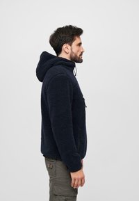 Sweat à capuche en polaire marine avec une surface texturée, poche avant et capuchon à cordon de réglage, porté avec un pantalon cargo olive. Vue de côté.