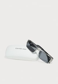 Ochelari de soare Calvin Klein Jeans cu lentile dreptunghiulare gri, cadru din metal argintiu și plastic negru, și un toc alb cu logo-ul brandului.