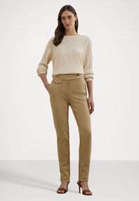 Krémový pletený sveter s textúrovaným vzorom, skombinovaný s béžovými nohavicami na mieru s detailom gombíkovej kapsy. Hnedé sandále na podpätku dopĺňajú celkový look.